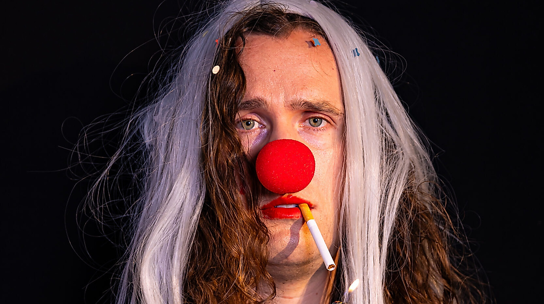 Eine Person im Clownkostüm hat eine Zigarette im Mund. Die Zigarette wird angezündet.
