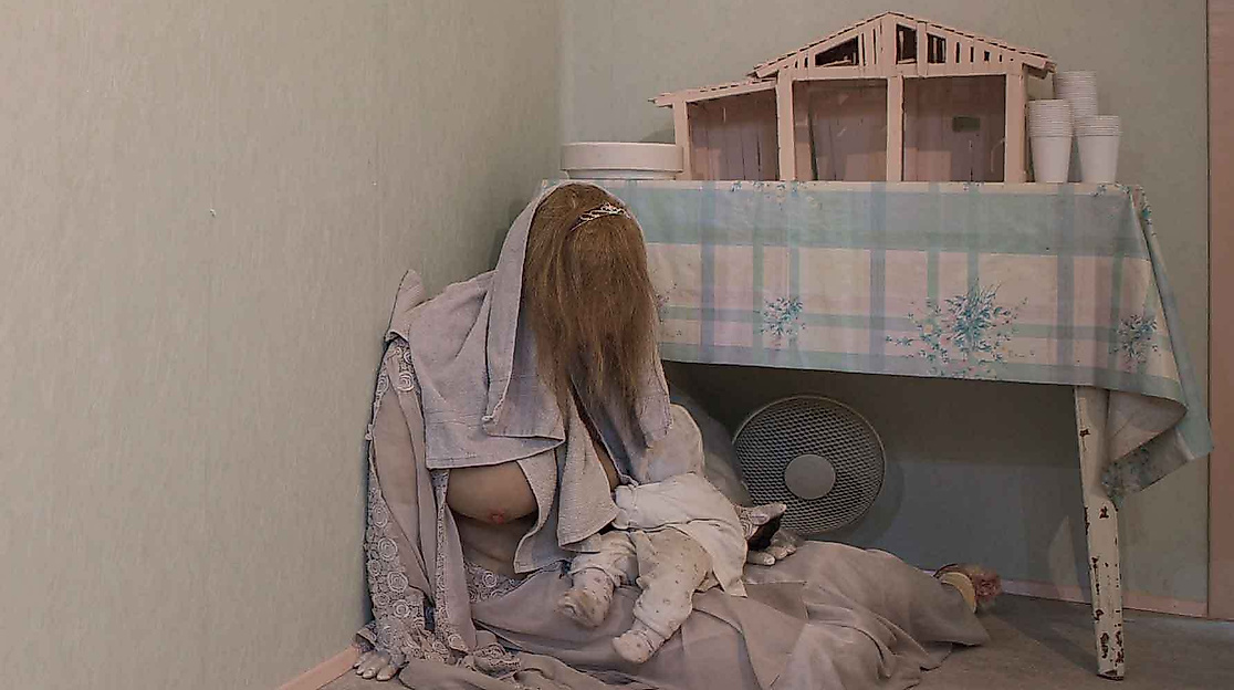 Eine Person mit langen Haaren sitzt mit einem Baby auf dem Boden neben einem Tisch mit einem Modellhaus.