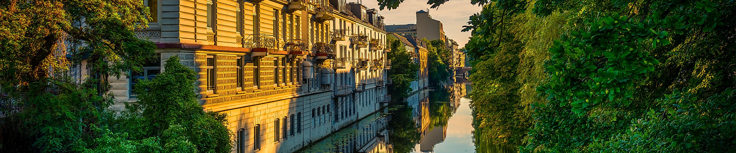 Jugendstilfassaden am Alsterkanal in Hamburg bei Sonnenuntergang, gesäumt von Bäumen und Wasserreflexionen