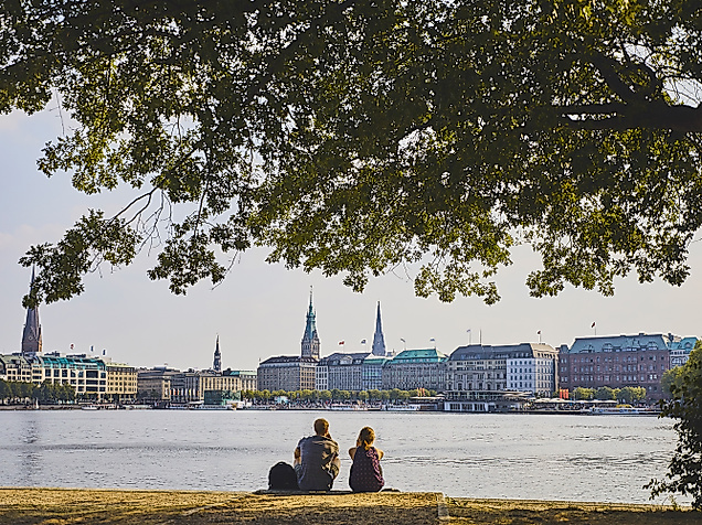 Zwei Personen sitzen am Alsterufer mit Blick auf Hamburgs Skyline aus Rathaus und Kirchtürmen im Sonnenlicht.