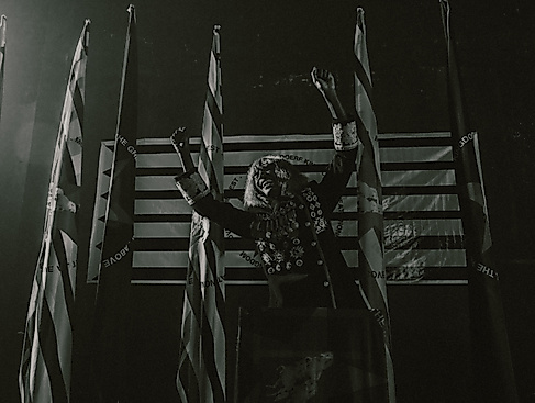 KABEAUSHÉ steht in einer Uniform mit vielen Orden auf einer Bühne vor vier Flaggen, streckt die Arme seitlich nach oben und lacht.