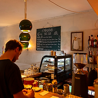 Gemütlicher Innenraum eines Cafés in Hamburg mit Theke, Kuchen­vitrine, warmem Licht und wartendem Gast