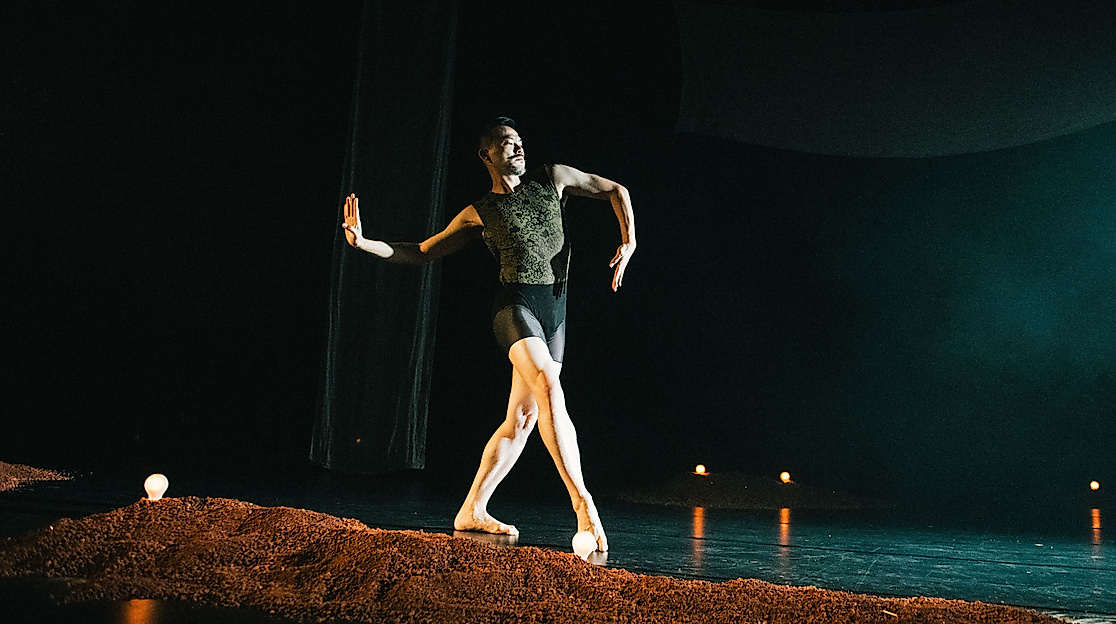Ausschnitt aus der Tanzproduktion »Choreographing Story »Rama House«« von Chen Wu-kang und Pichet Klunchun. Eine performende Person bewegt sich in einer Ballett-Pose über die Bühne. Vor ihr ist ein kleiner erdartiger Hügel aufgescharrt.