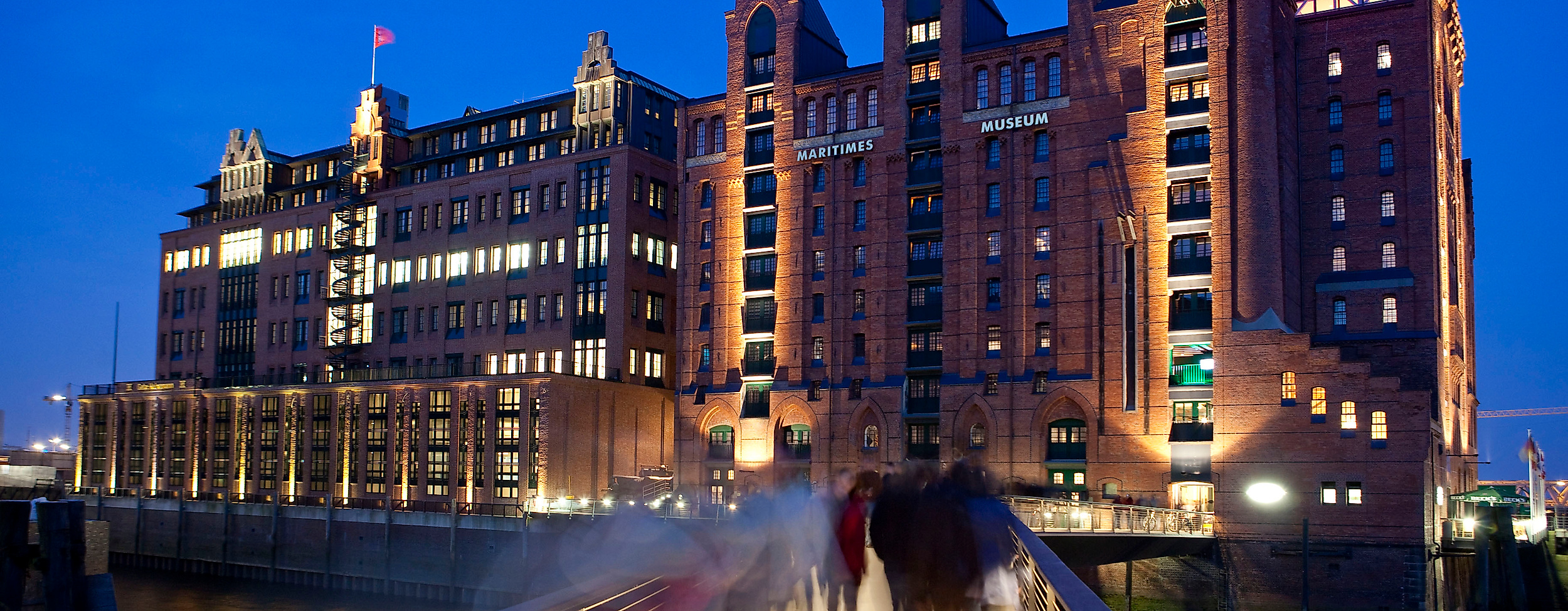 Internationales Maritimes Museum in der beleuchteten Speicherstadt am Abend mit Fußgängerbrücke im Vordergrund.