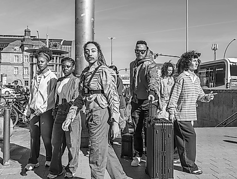 Sieben Menschen beim verlassen einer U-Bahnstation. Vier von ihnen schauen ernst in die Kamera, während drei im Hintergrund zur Seite laufen.