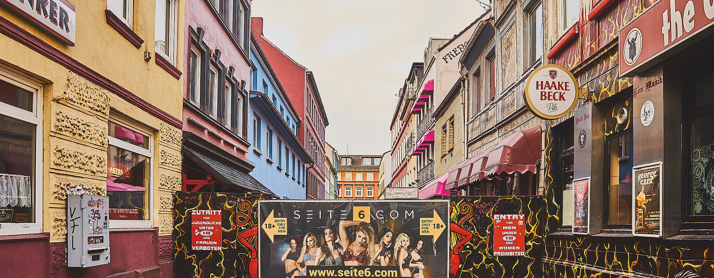 Blick in die Herbertstraße mit Sichtschutzwand, flankiert von bunt beleuchteten Fassaden und Schildern auf der Reeperbahn