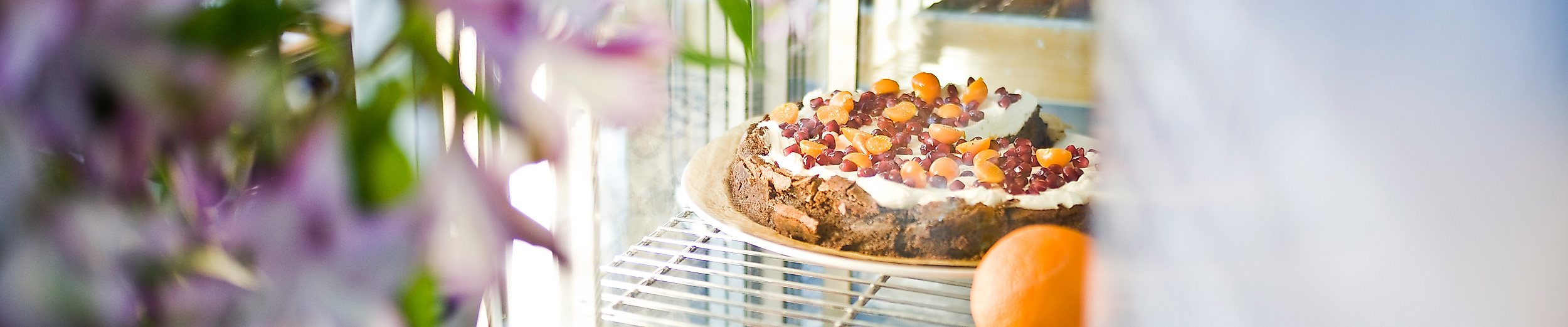 Kuchenvitrine mit frischen Torten und Orangen im Café Johanna, daneben Frühlingsblumen in einer Vase