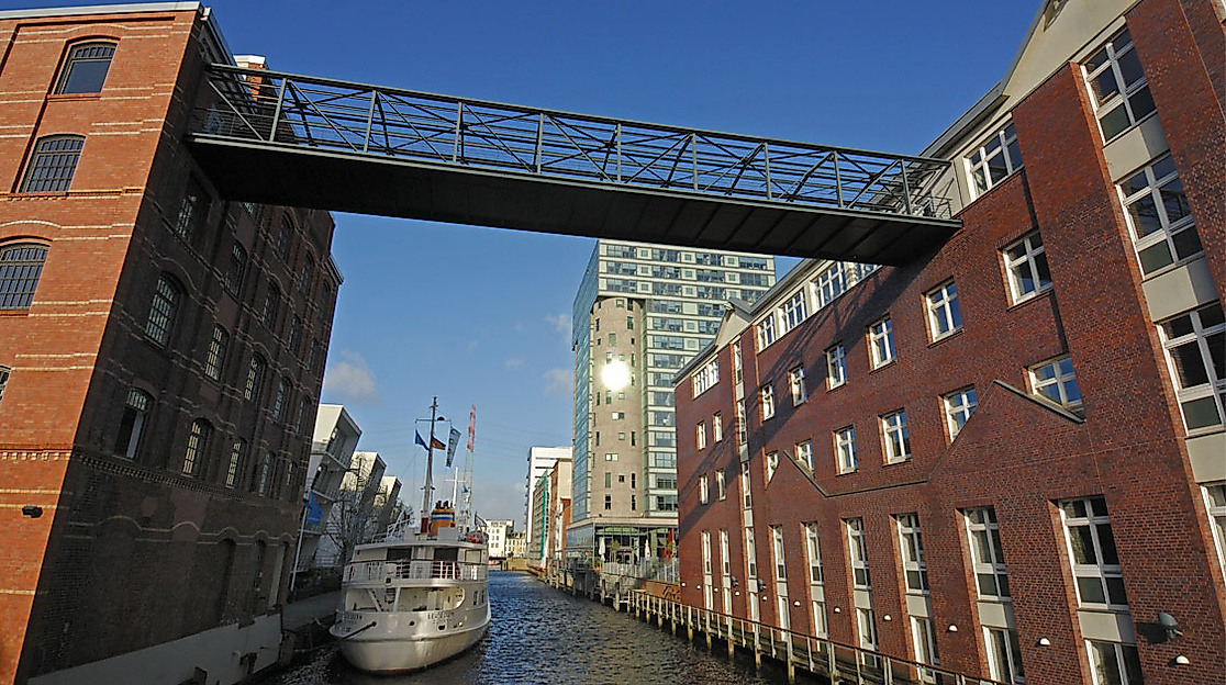 Backsteinbauten am Kanal in Hamburg Harburg mit moderner Fußgängerbrücke und vorbeifahrendem Boot bei Sonne.