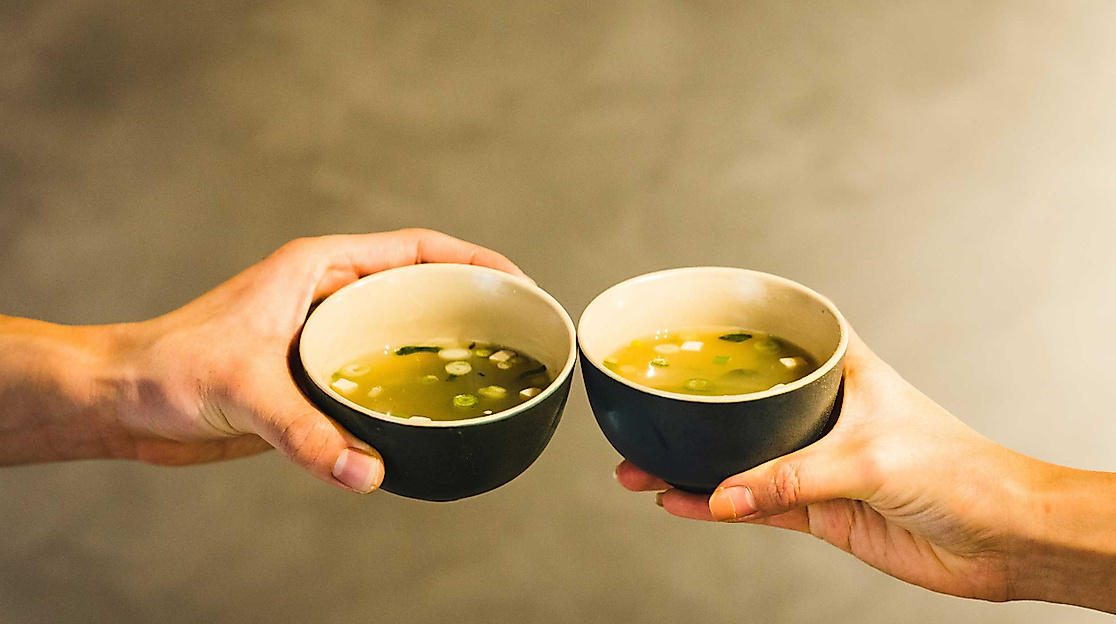 Zwei Hände stoßen mit dampfenden Schalen Ramen-Suppe vor neutralem Hintergrund an