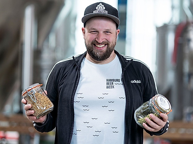 Lächelnder Mann mit Cap hält zwei Gläser mit Brauzutaten in Brauerei bei der Hamburg Beer Week