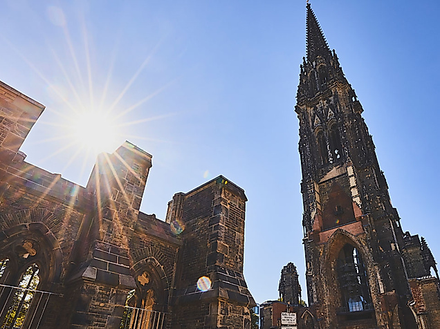 Ruine der St.-Nikolai-Kirche in Hamburg bei Sonnenschein, mit Mahnmal und neugotischem Turm vor blauem Himmel
