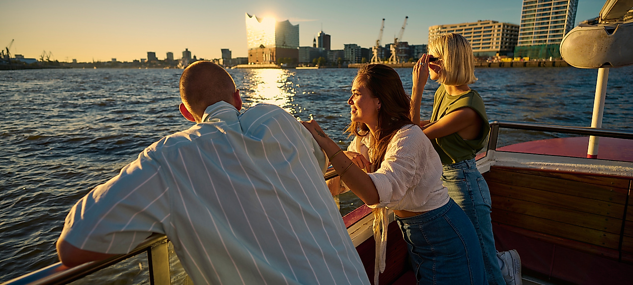 Junge Erwachsene auf einem Boot auf der Elbe. Im Hintergrund ist die Elphi zu sehen.
