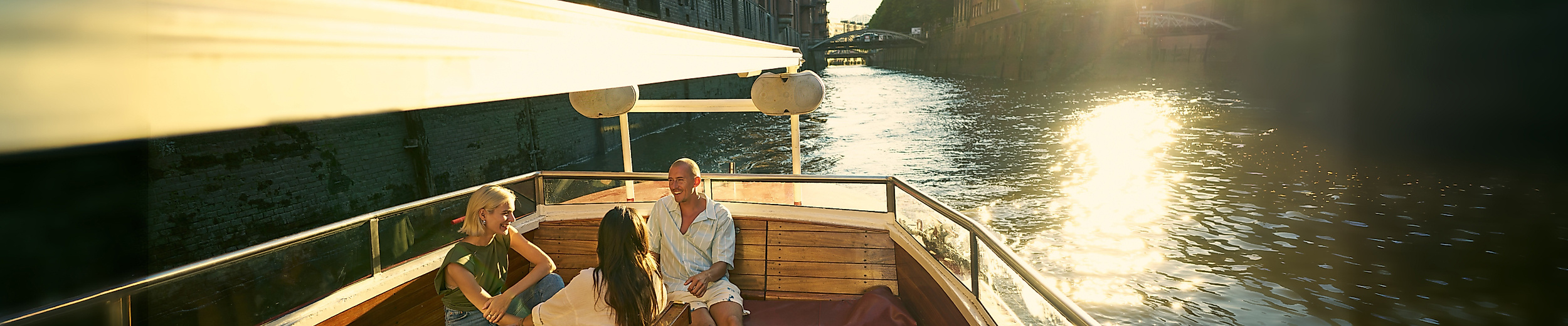 Drei Personen auf einer Barkasse bei Sonnenuntergang mit der Speicherstadt im Hintergrund