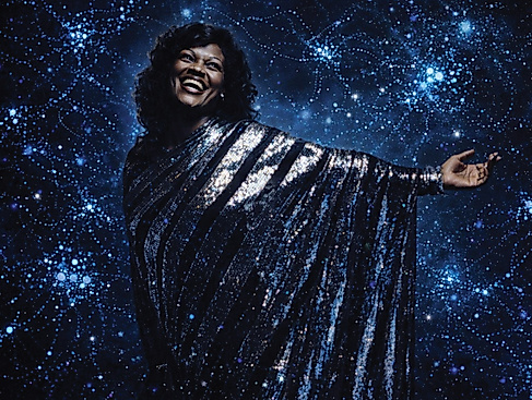 Eine Frau steht in einem silber-glitzernden Kleid vor dem Hintergrund einer dunkelblauen Fläche, auf der viele Lichter sind.