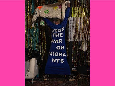 Hools of Fashion: Eine Person steht mit dem Rücken zugewandt vor einer Wand voller lamettaartigen Strukturen. Auf ihrem Kleid steht »Stop the War on Migrants«.