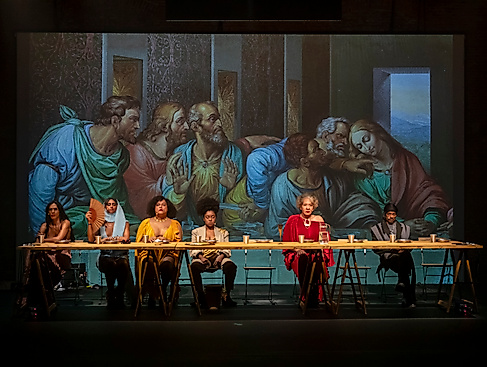 Performer*innen sitzen an einer langen Tafel vor einer Projektion von Da Vincis »Letztem Abendmahl« — eine zeitgenössische Inszenierung mit vielfältigem Ensemble.