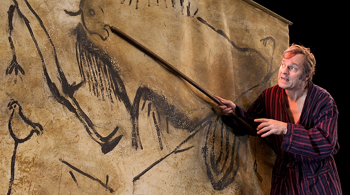 Szenenbild Cavemen: Ein Mann in Bademantel zeigt mit einem Stock auf eine große Wand mit Zeichnungen