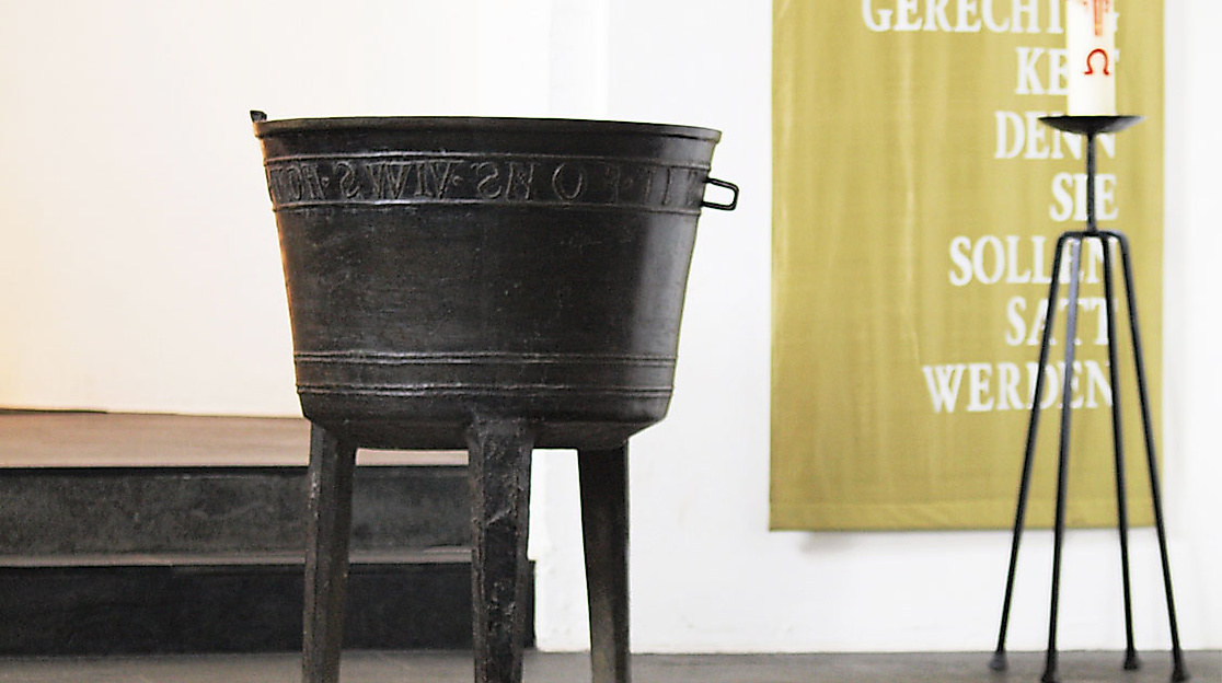 Historisches Taufbecken in der Kirche Blankenese vor hellem Hintergrund mit Kerzenständer und Wandbehang