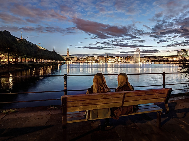 Zwei Personen sitzen bei Abenddämmerung an der Binnenalster mit Blick auf die beleuchtete Stadt