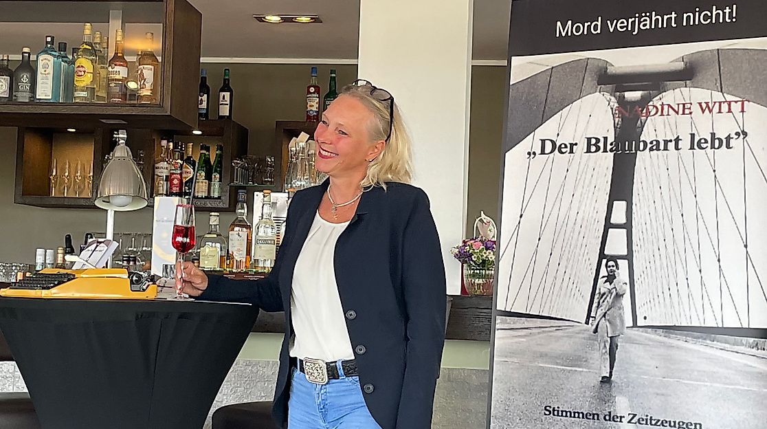 Die Autorin Nadine Witt steht neben einem Roll-up mit dem Cover ihres Buches „Der Blaubart lebt“ – Stimmen der Zeitzeugen. Sie blickt leicht zur Seite und trägt einen Ausdruck positiver Ausstrahlung im Gesicht. Das Roll-up zeigt das düstere Buchmotiv. Witt bezeichnet es selbst als „Das zweite Kapitel einer düsteren Legende“.