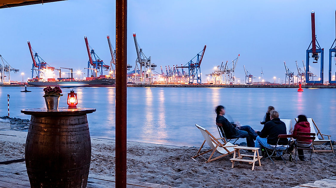 Abendstimmung im Strandrestaurant Strandperle mit Blick auf den beleuchteten Hamburger Hafen und sitzende Gäste am Elbstrand