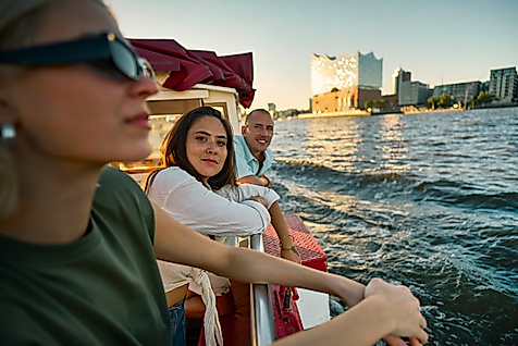 Drei junge Menschen auf einem Boot auf der Elbe. Im Hintergrund ist die Elbphilharmonie.