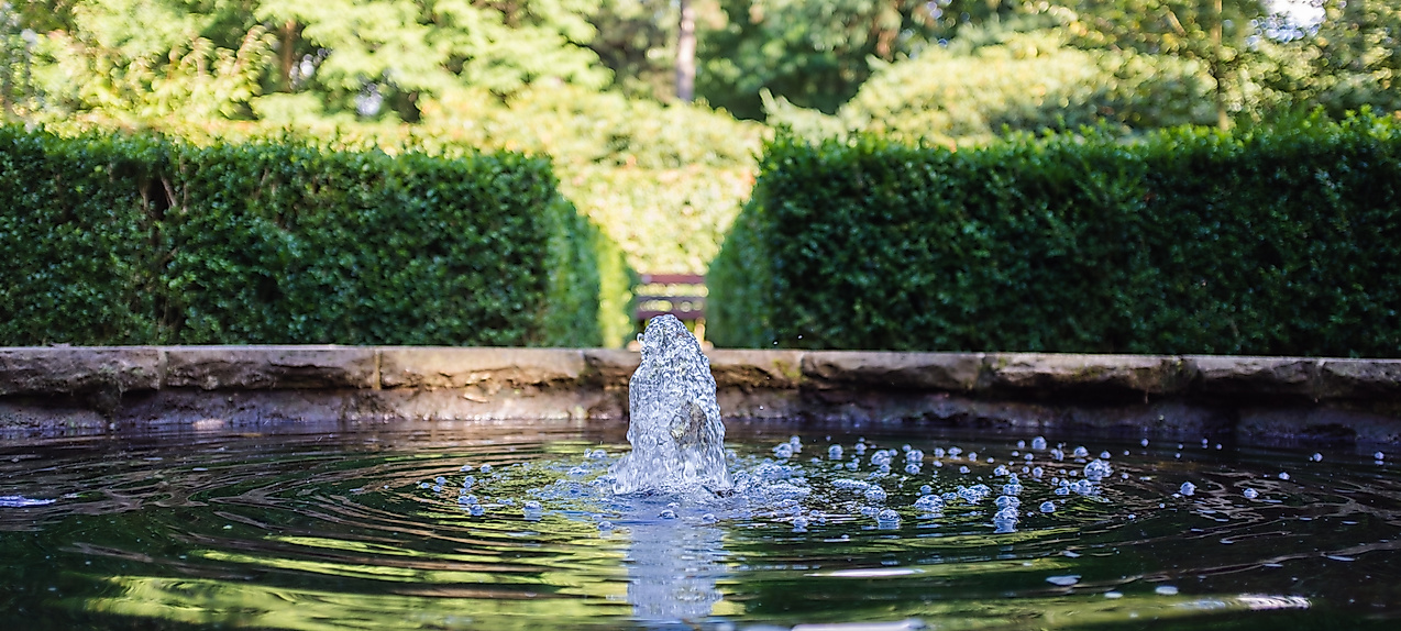 Wasser sprudelt aus einem Brunnenbecken, eingefasst von Hecken und Bäumen in einer ruhigen Parkanlage.