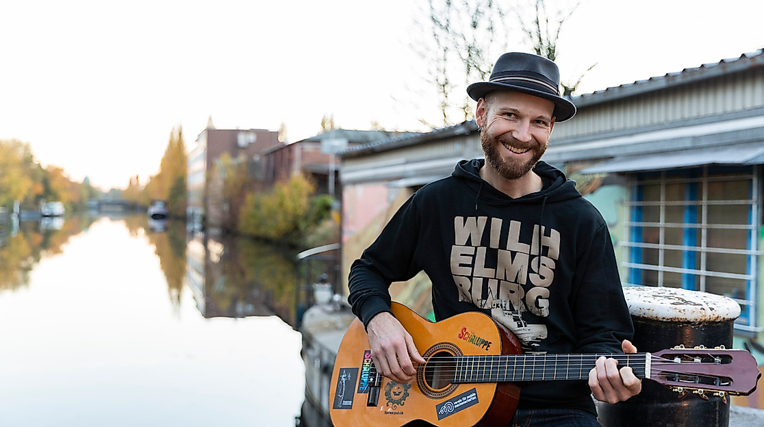 Mann mit Gitarre und Hut sitzt am Wasser vor Hausboot in Hamburg, lächelt in die Kamera bei Herbstlicht