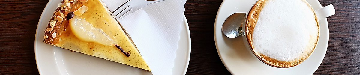 Stück Birnentarte mit Puderzucker und Cappuccino auf Holztisch, serviert in der Biokonditorei Eichel