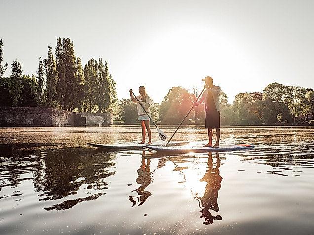 Zwei Personen beim Stand-up-Paddling im Gegenlicht auf ruhigem Wasser vor Bäumen und Abendsonne