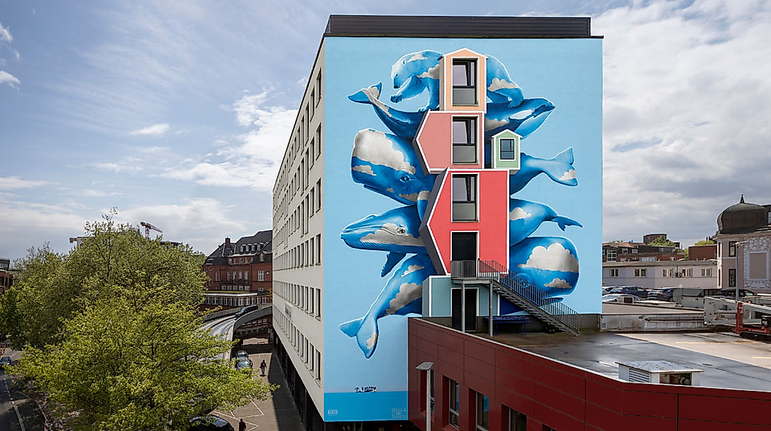 Bunte Hausfassade in Hamburg-Harburg mit 3D-Wandgemälde fliegender Fische auf blauem Hintergrund bei Sonnenschein.