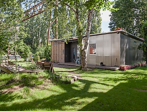 Foto vom Migrantpolitan auf Kampnagel. Ein kleines einstöckiges Haus mit einem grünen Garten, Bänken aus Holzpaletten, umsäumt von vielen Bäumen.