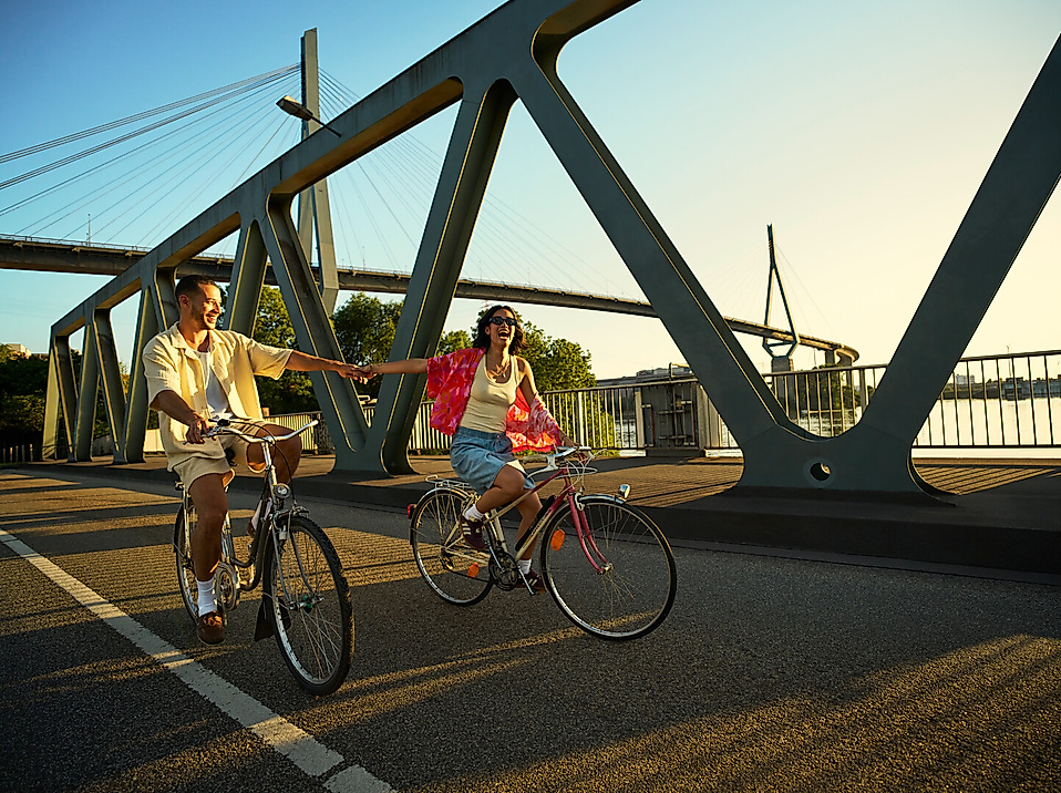Zwei junge Leute fahren lachend mit dem Fahrrad über eine Brücke und halten sich dabei an den Händen.