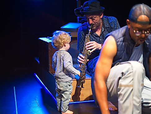 Ein männlich-gelesener Musiker mit einer Klarinette spielt während er seinen Blick einem kleinen Kind zuwendet. Dieses steht der Klarinette zugewandt vor ihm. Im Vordergrund ist verschwommen ein weiterer Musiker zu sehen.