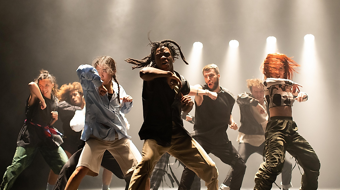 Eine energiegeladene Tanzgruppe führt auf einer Bühne unter dramatischer Beleuchtung eine kraftvolle Choreografie auf.