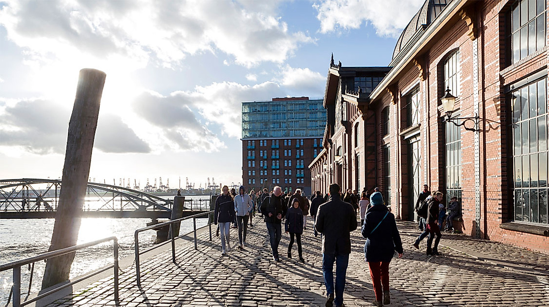 Spaziergänger:innen an der Hamburger Fischauktionshalle bei Sonnenschein mit Blick auf die Elbe