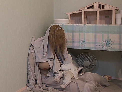 Eine Person mit langen Haaren sitzt mit einem Baby auf dem Boden neben einem Tisch mit einem Modellhaus.