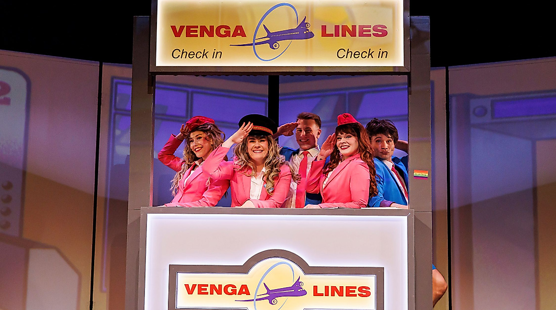 Szenenbild Tschüssikowski: Fünf Personen in pink-blauen Uniformen stehen in Nachbildung eines Flugzeugfensters mit Aufschrift „Venus Air“.