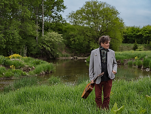 Ron Sexsmith steht in einer grünen Landschaft und hält seine Gitarre in der Hand.