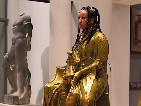 Eine Person in einen glänzenden goldfarbenen Anzug sitzt als Ausstellungsstück im Museum.