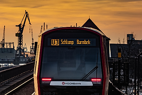 U-Bahn der Linie U3 Richtung Barmbek bei Sonnenuntergang, mit Hafenkränen im Hintergrund.