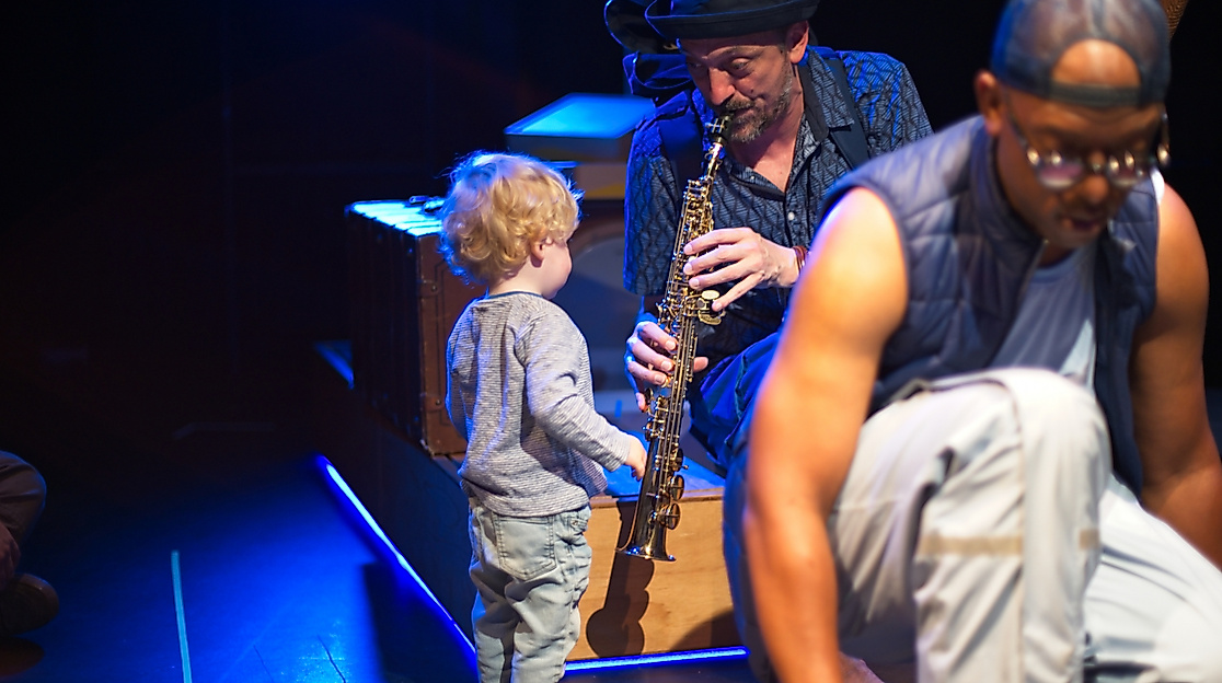 Ein männlich-gelesener Musiker mit einer Klarinette spielt während er seinen Blick einem kleinen Kind zuwendet. Dieses steht der Klarinette zugewandt vor ihm. Im Vordergrund ist verschwommen ein weiterer Musiker zu sehen.